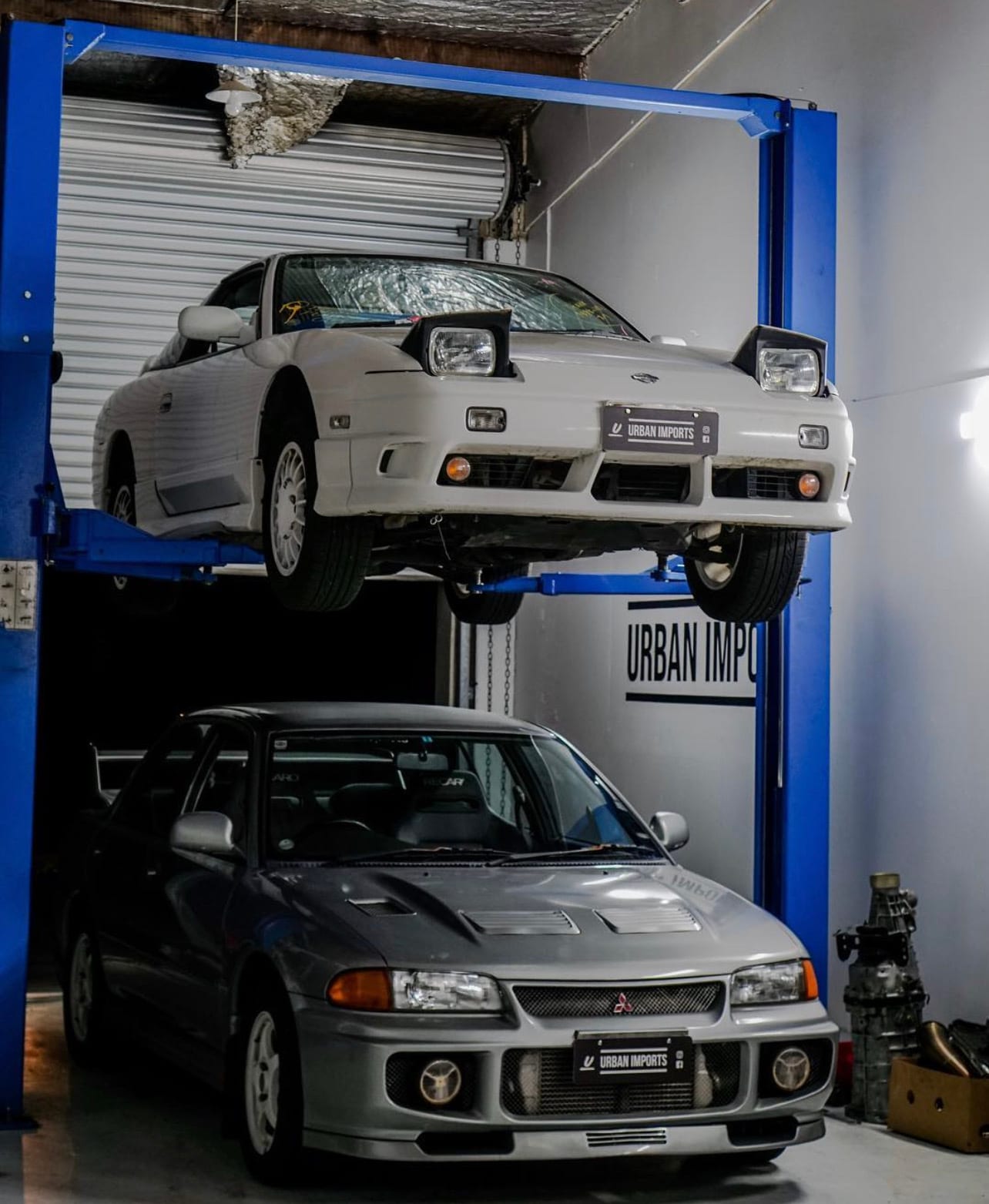 A Nissan Skyline raised on the hoist above a Lancer Evolution in the Urban Imports workshop